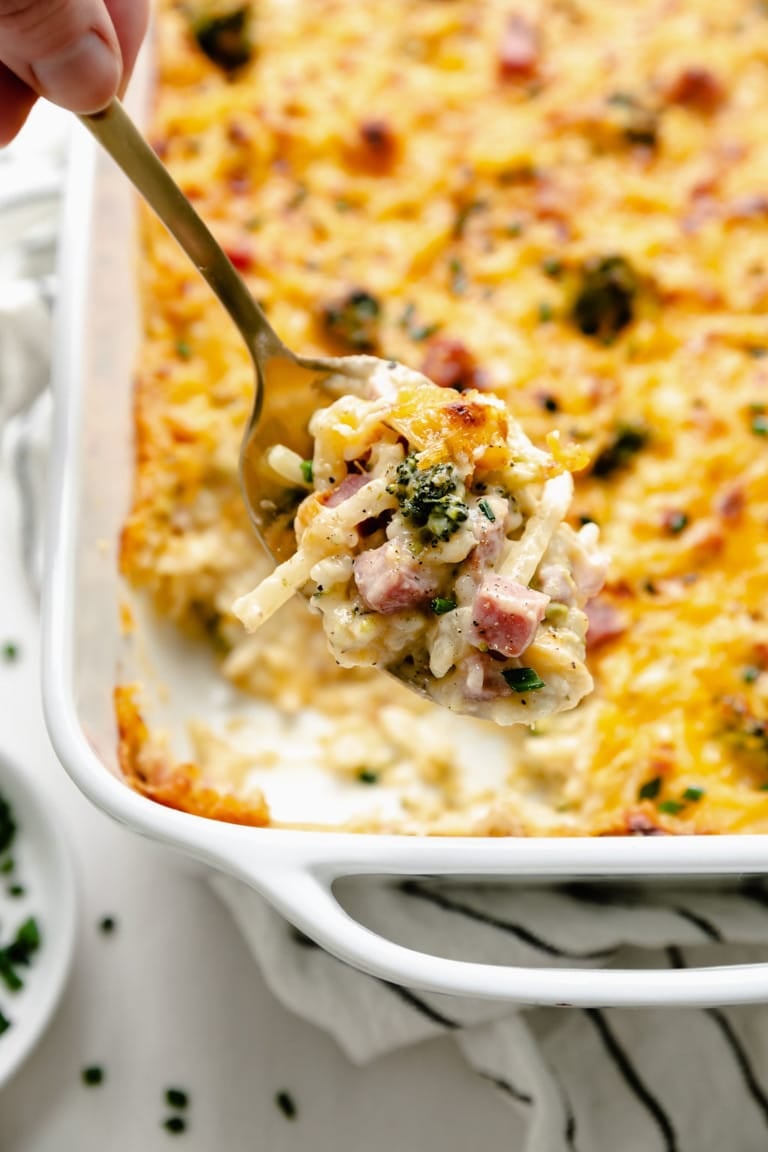 A scoop being taken from a freshly baked pan of cheesy scalloped potatoes with broccoli and ham 