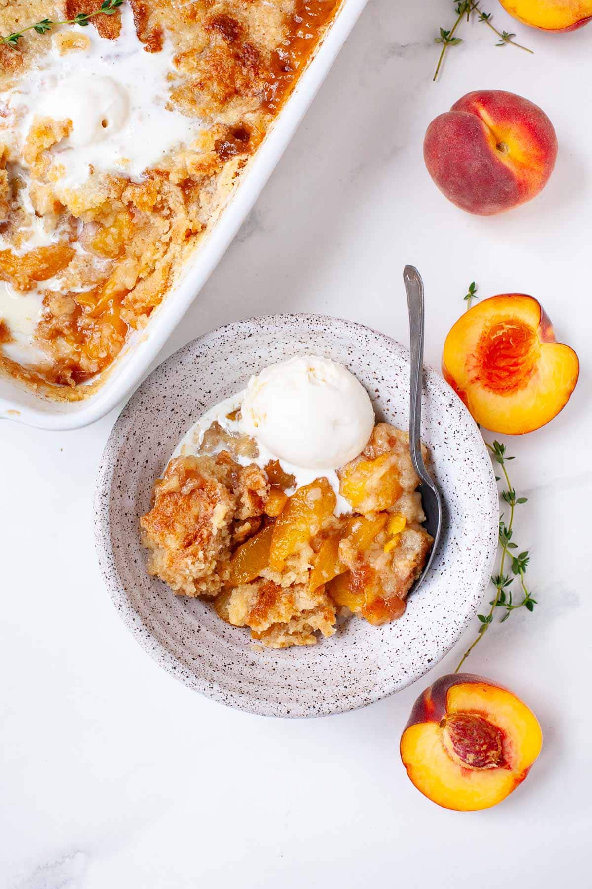 cake mix peach cobbler with scoop of vanilla ice cream in white speckled bowl on white marble surface with whole and sliced peaches