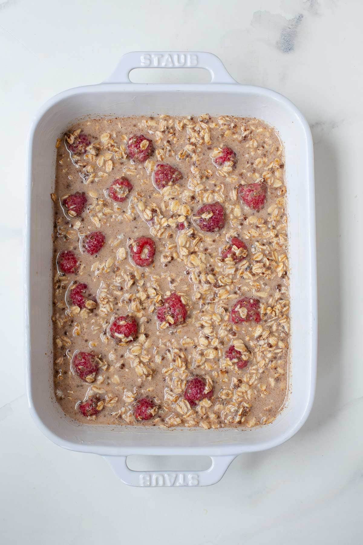 Prepped vegan oatmeal mixture with raspberries in white ceramic baking dish on marble counter.