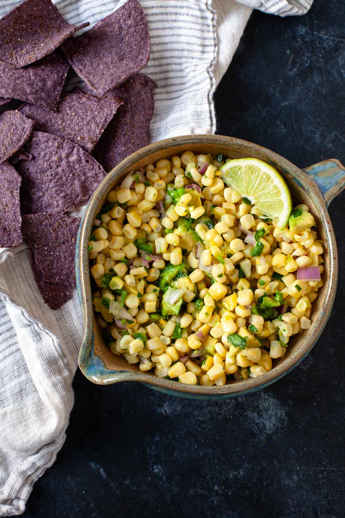 corn avocado salsa with lime wedge in brown and blue ceramic bowl next to blue tortilla chips on grey striped dish towel