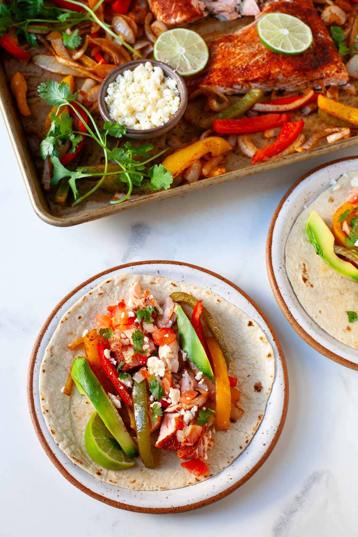 flaky salmon with fajita veggies on a tortilla next to a sheet pan of the ingredients